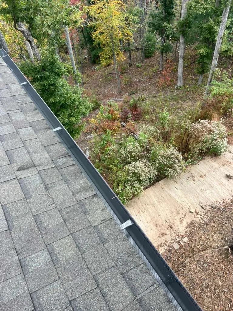 Eave fitted with a mesh gutter guard, the ground falling away into a wooded hillside below