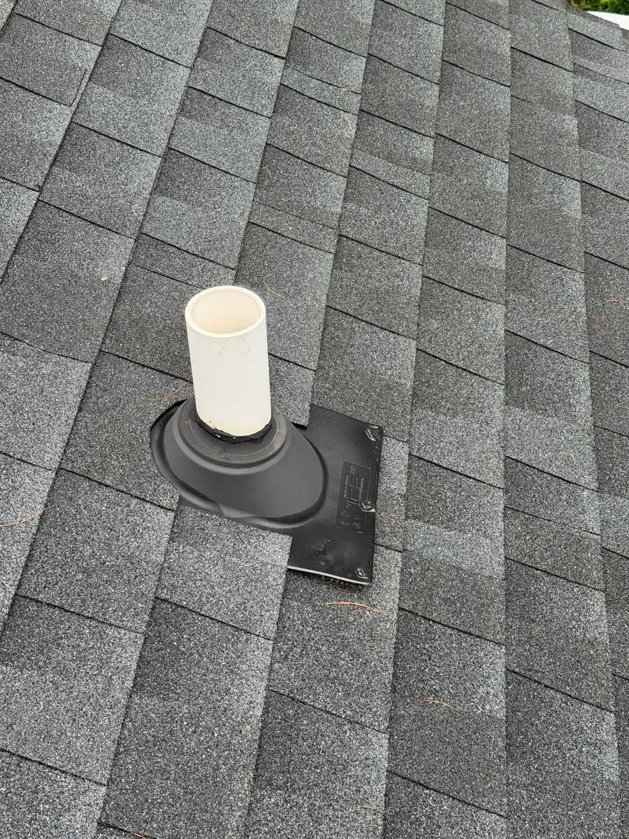 White PVC plumbing vent through a new rubber boot and its flashing plate on dark grey shingles