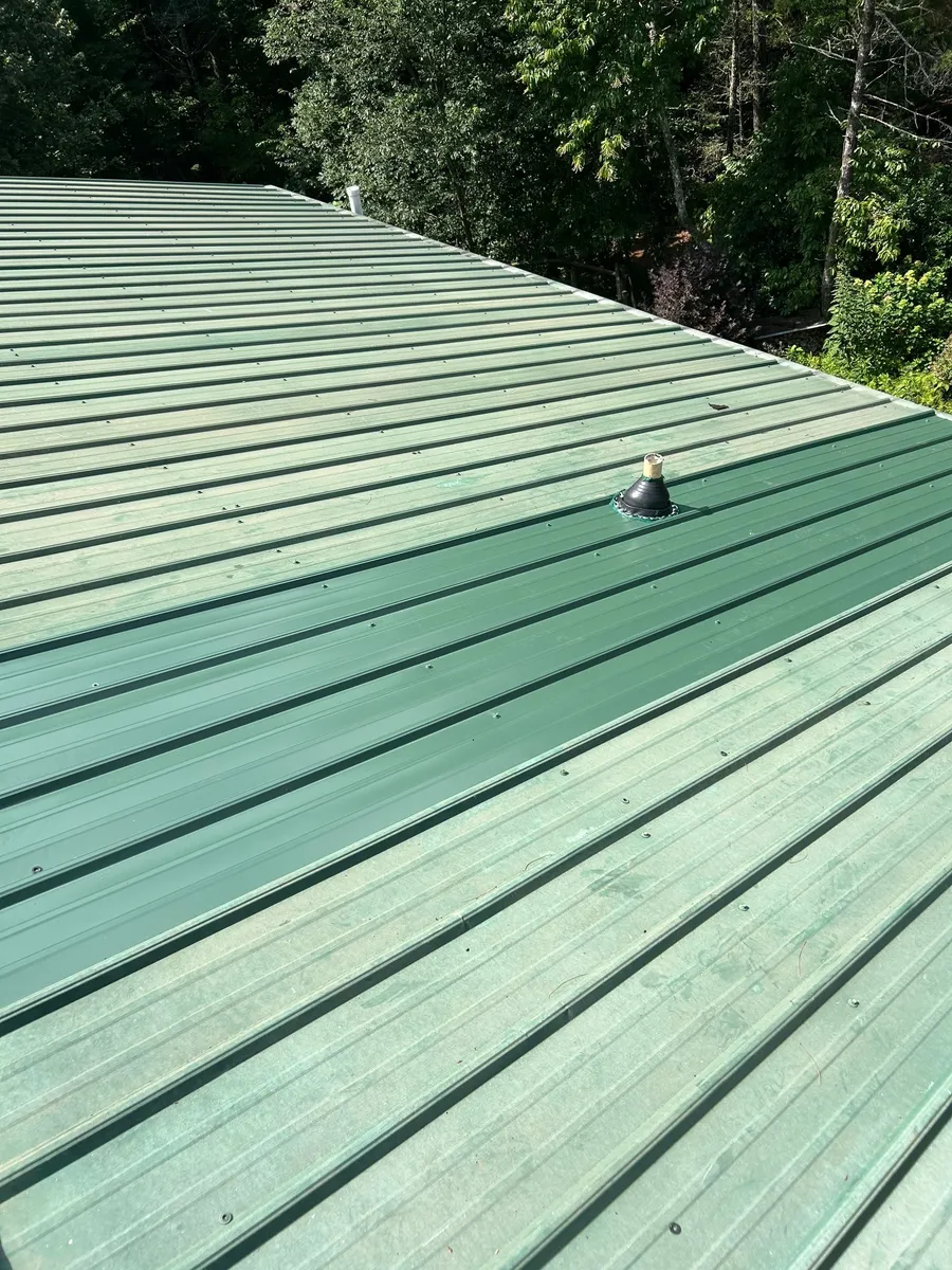 Long run of green standing-seam metal roofing with a sealed pipe penetration mid-slope, hardwoods behind