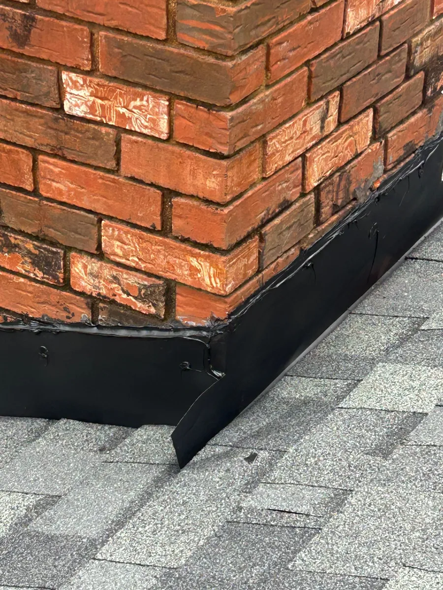 Black base flashing wrapped tight to a red brick chimney where it meets new grey shingles